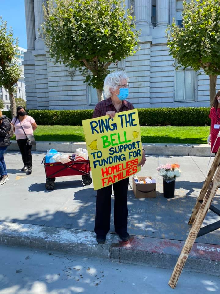 An older woman stands with a face mask and a sign that reads "ring the bell to support funding for homeless families"