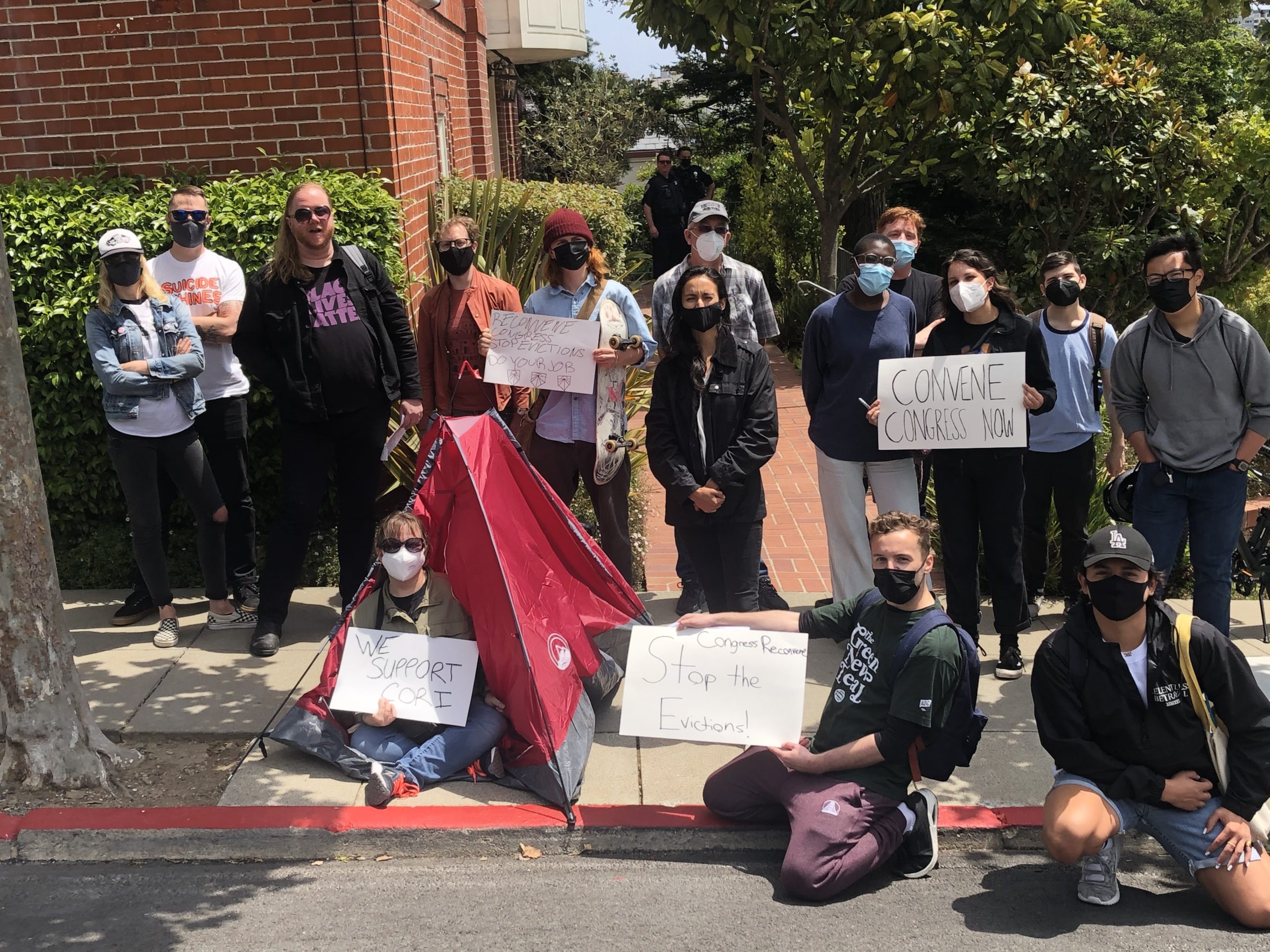 A group of protestors gathered outside Nancy Pelosi's mansion, a tent visible. They hold signs supporting Rep. Cori Bush